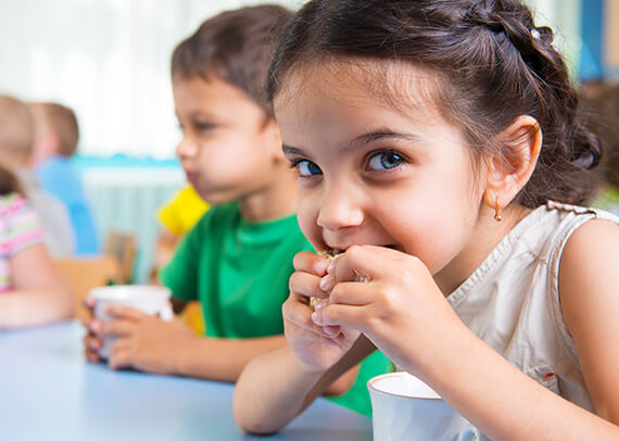 Young girl eating a snack with other kids sitting at the same table in the background.